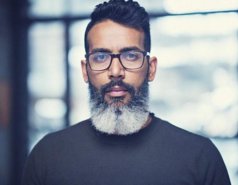 Mann mit Brille und grauem Bart, freundlich lächelnd, vor unscharfem Hintergrund.